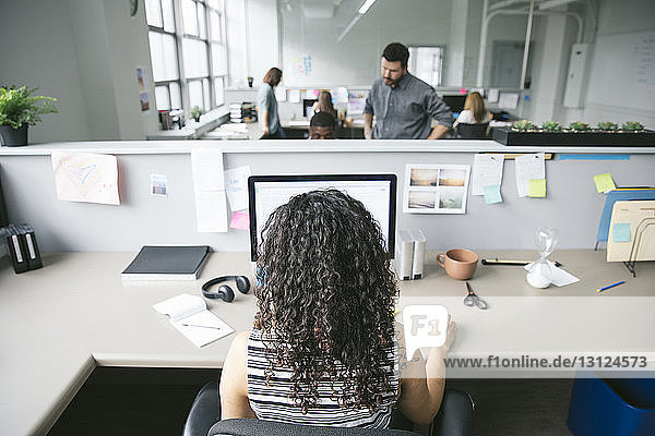 Rückansicht einer Geschäftsfrau  die am Desktop-Computer mit Kollegen im Hintergrund arbeitet