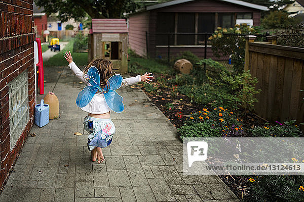 Rückansicht eines verspielten Mädchens mit Kostümflügeln beim Laufen im Hinterhof