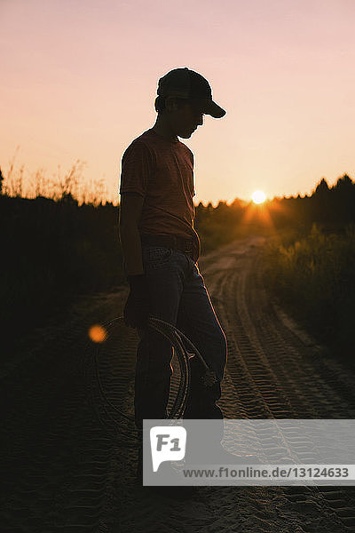 Full length of boy wearing cap while holding rope on dirt road against clear sky during sunset