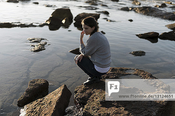Thoughtful woman looking down while crouching at rocky beach during sunset