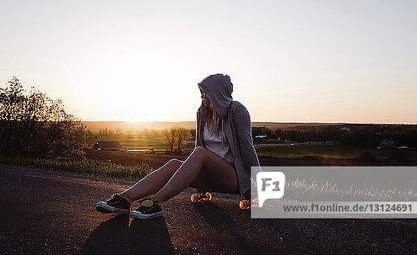 Frau mit Skateboard sitzt bei Sonnenuntergang bei klarem Himmel auf der Straße