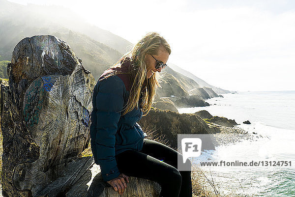Seitenansicht einer glücklichen Frau  die auf einem Felsen am Meer sitzt