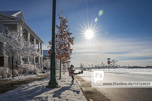 Seitenansicht eines verspielten Mädchens  das bei Sonnenschein Schnee auf der Straße gegen den blauen Himmel tritt