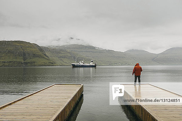 Rückansicht eines Mannes in Kapuzenjacke  der bei nebligem Wetter auf dem Pier gegen die Berge steht