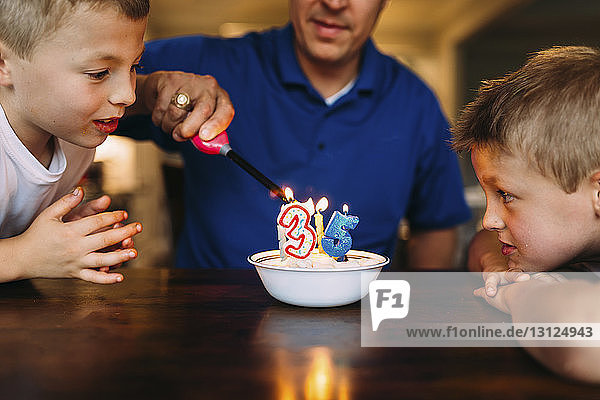 Neugierige Brüder sehen Vater beim Anzünden von Geburtstagskerzen mit Feuerzeug auf dem Tisch zu Hause