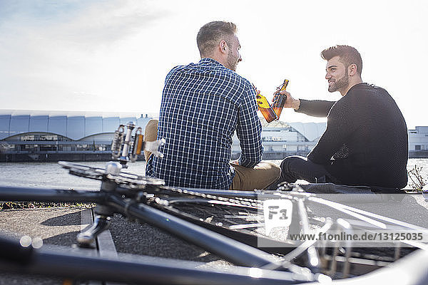 Friends toasting beer bottle while sitting by bicycle on promenade against clear sky