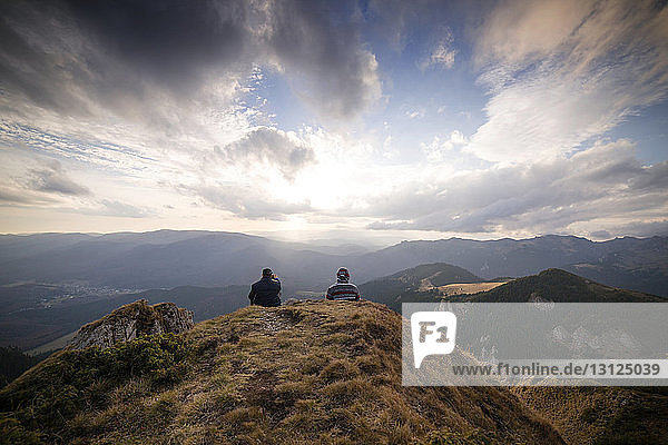 Rear view of men sitting on mountain against cloudy sky