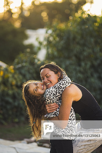 Mother with eyes closed embracing daughter while standing in yard during sunset