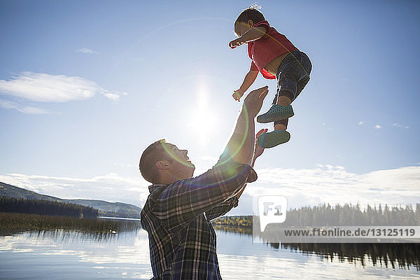 Side view of playful father throwing son in air against lake and sky during sunny day