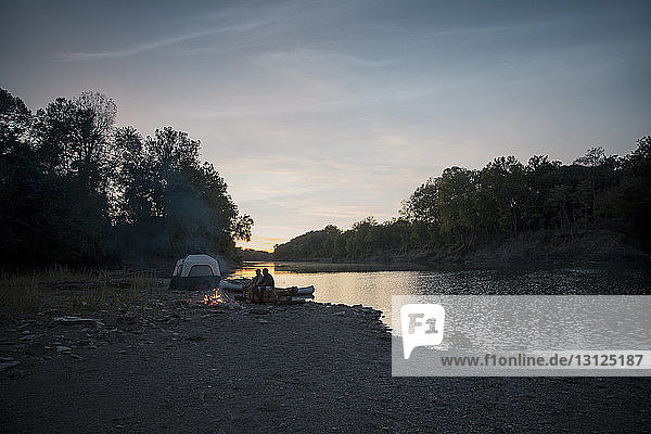 Männliche Freunde sitzen bei Sonnenuntergang auf einem Baumstamm auf einem Campingplatz am See gegen den Himmel
