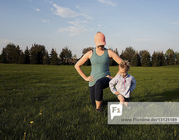 Mother with daughter exercising on grassy field at park against sky