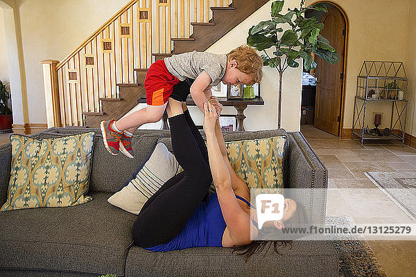 Seitenansicht einer spielerischen Mutter  die ihren Sohn im heimischen Wohnzimmer auf den Beinen balanciert