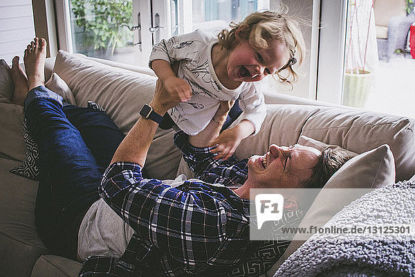Happy father lifting up daughter while lying on sofa at home