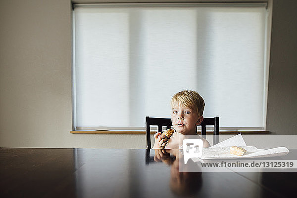 Portrait of shirtless boy eating bread while sitting on chair at home