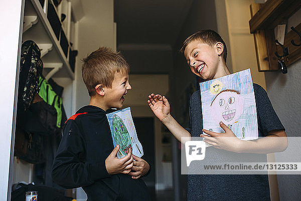 Fröhliche Brüder halten Zeichnungen in der Hand  während sie zu Hause stehen