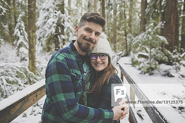 Portrait of smiling couple embracing on footbridge amidst trees in forest at Lynn Canyon Park during winter