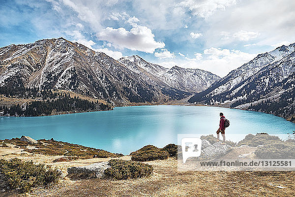 Full length of backpacker looking at view while standing by lake against mountains and cloudy sky during winter