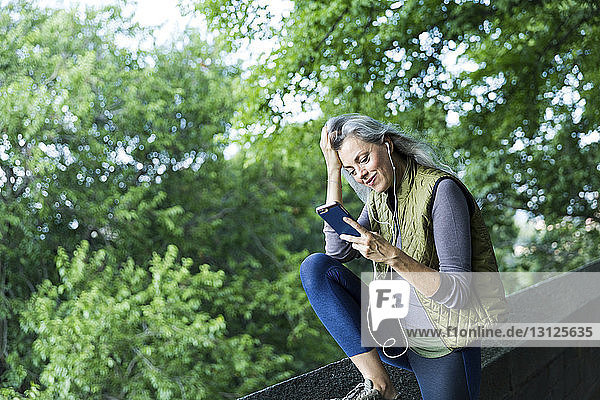 Woman with hand in hair using smart phone while listening music by retaining wall against branches