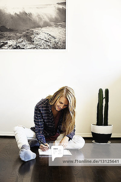 Lächelnde Frau schreibt in Buch  während sie zu Hause auf dem Boden sitzt