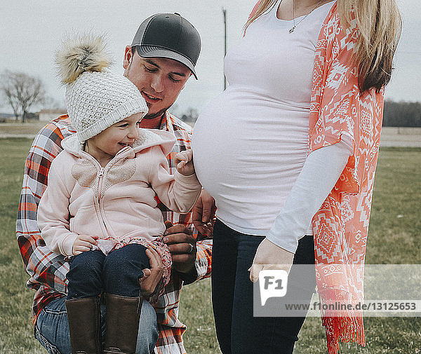 Vater trägt fröhliche Tochter von schwangerer Frau auf Feld im öffentlichen Park