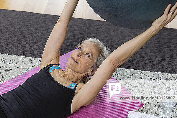 Ältere Frau hält Fitnessball in der Hand während sie zu Hause auf einer Trainingsmatte liegt