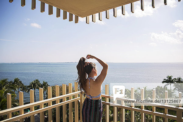 Rückansicht einer Frau  die sich die Haare bindet  während sie auf dem Balkon gegen das Meer steht