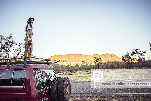 Portrait of smiling woman with camera standing on car roof at desert