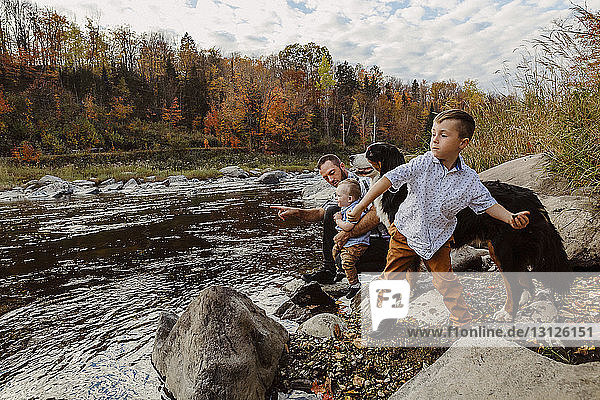 Father with sons and dog enjoying by river against cloudy sky in forest