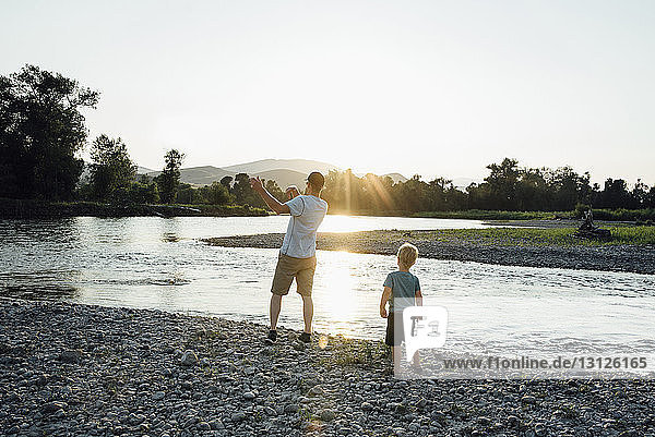 Rückansicht eines Vaters mit Sohn beim Spielen am Seeufer vor klarem Himmel bei Sonnenuntergang