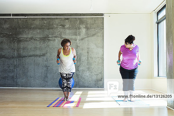 Full length of woman looking at friend standing on exercise mat while holding dumbbells against wall in yoga studio