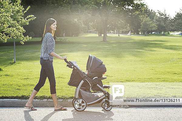 Side view of smiling woman walking with baby stroller on street at park