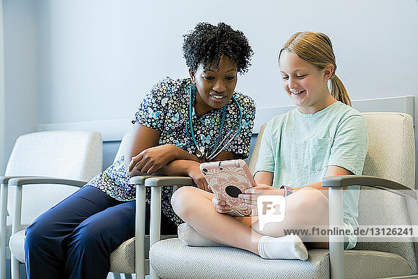 Kinderarzt betrachtet Mädchen mit Tablet-Computer  während sie im Krankenhaus auf einem Stuhl sitzt