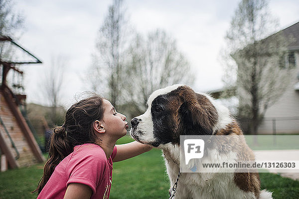 Glückliches Mädchen küsst Hund  während es auf einem Grasfeld gegen den Himmel im Hof sitzt
