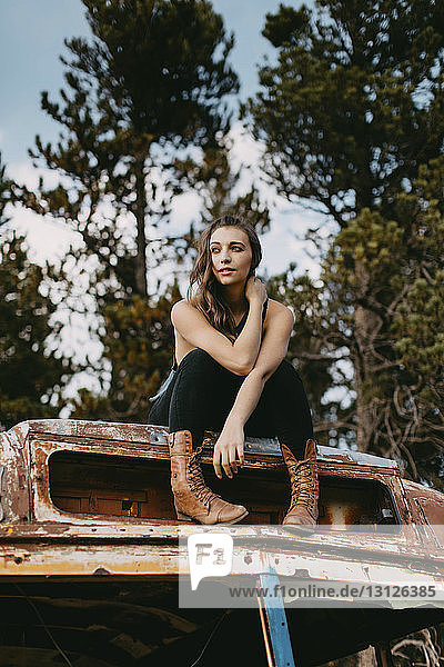 Niedrigwinkelansicht einer nachdenklichen jungen Frau  die weg schaut  während sie auf einem verlassenen Fahrzeug im Wald sitzt