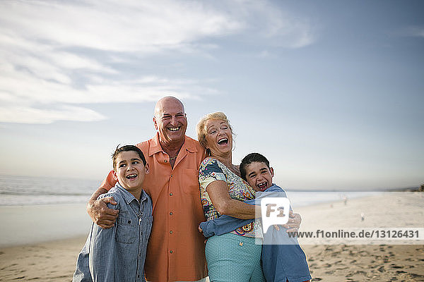 Happy grandparents with grandsons standing at beach against sky