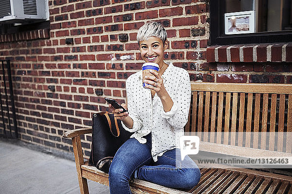 Porträt einer Frau  die auf einer Bank sitzend ein Mobiltelefon und eine Kaffeetasse hält