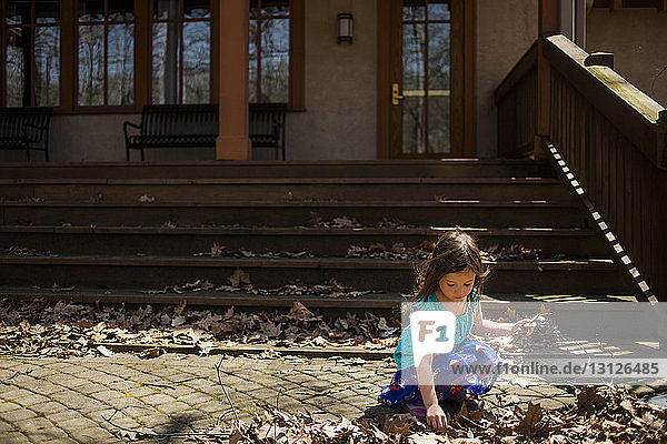 Mädchen sammelt trockenes Laub  das im Herbst auf einem Fußweg gegen ein Haus gefallen ist