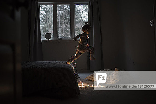 Seitenansicht eines Mädchens mit beleuchteten Lichterketten  das zu Hause vom Bett springt