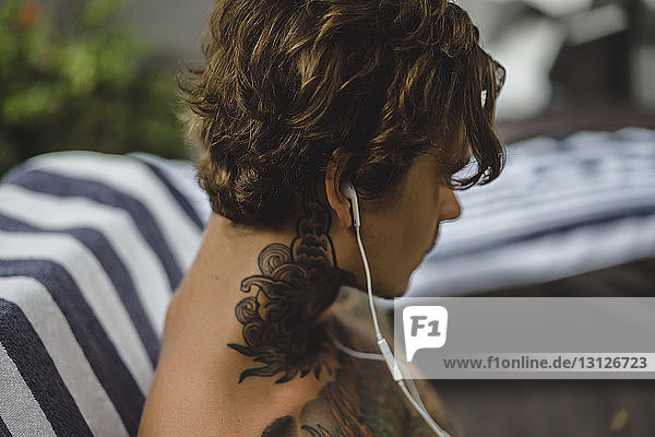 Nahaufnahme eines ohne Hemd tätowierten Mannes  der zu Hause über Kopfhörer Musik hört