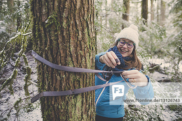 Lächelnde Frau bindet im Wald im Lynn Canyon Park im Winter Hängemattenbänder an Baumstamm