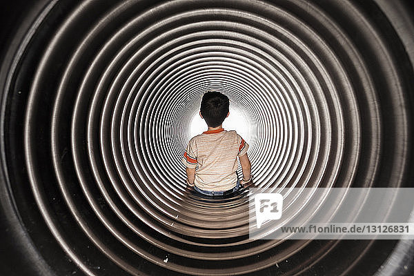 Rear view of boy sitting in huge metallic pipe