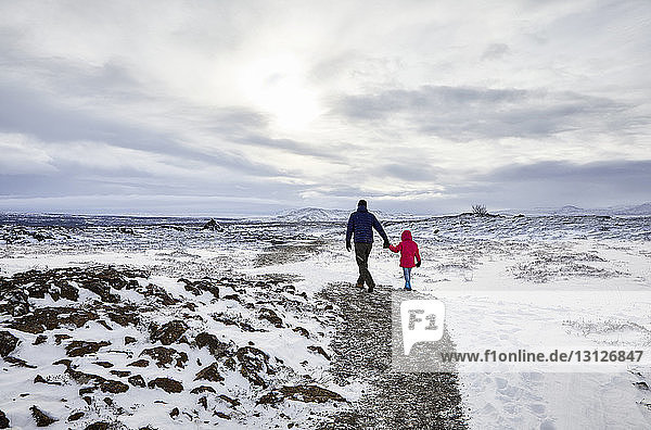 Rückansicht eines Vaters und einer Tochter  die sich an den Händen halten  während sie über eine schneebedeckte Landschaft vor bewölktem Himmel gehen