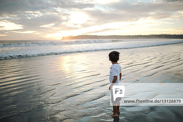Side view of boy standing at beach against cloudy sky during sunset
