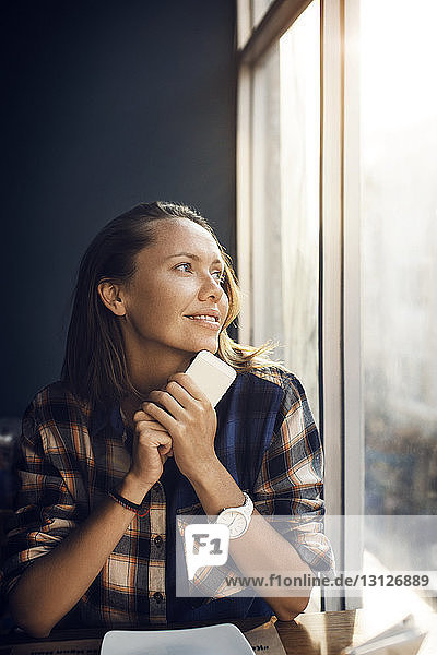 Lächelnde Frau hält Smartphone in der Hand  während sie durchs Fenster in ein Café schaut