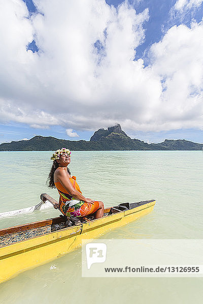 Frau in traditioneller Kleidung beim Kanufahren auf der Lagune der Insel Bora Bora vor bewölktem Himmel