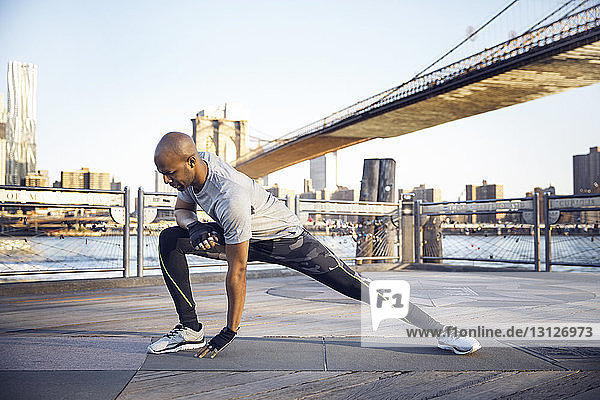 Männlicher Athlet in voller Länge beim Training auf der Promenade mit der Brooklyn Bridge im Hintergrund