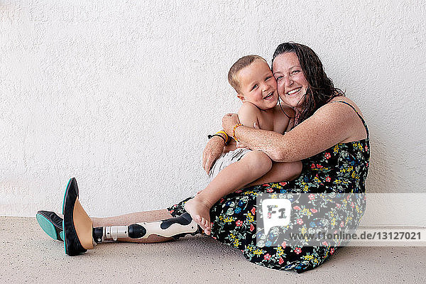 Portrait of mother with prosthetic leg embracing son while sitting against wall