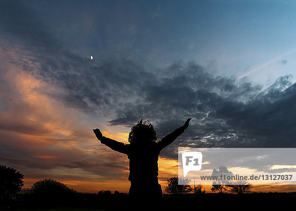 Silhouette eines fröhlichen Jungen mit erhobenen Armen vor bewölktem Himmel stehend