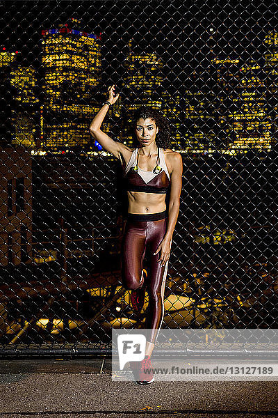 Portrait of female athlete standing by fence against illuminated buildings at night