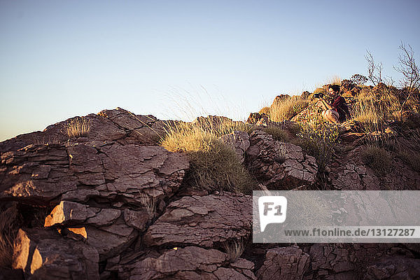 Low angle view of woman photographing while sitting on rock formations against clear sky